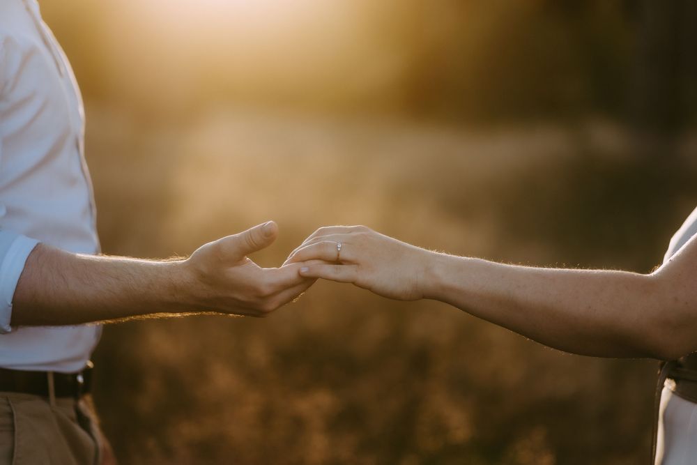 Close-up of couple holding hands with engagement ring in warm golden sunset light outdoors.