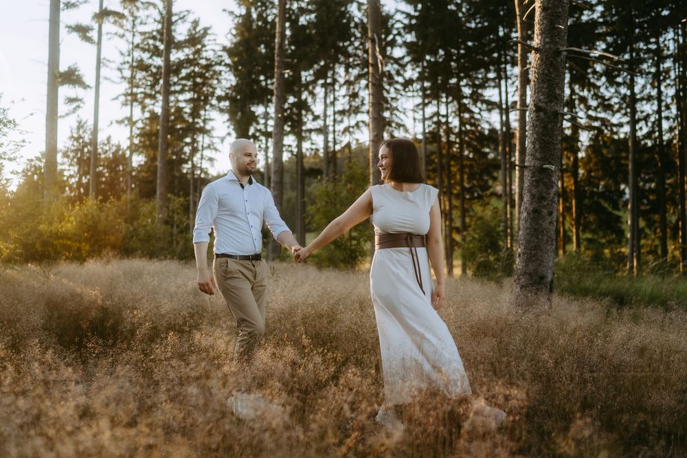 Couple holding hands walking through golden meadow with tall pine trees in warm sunset light.