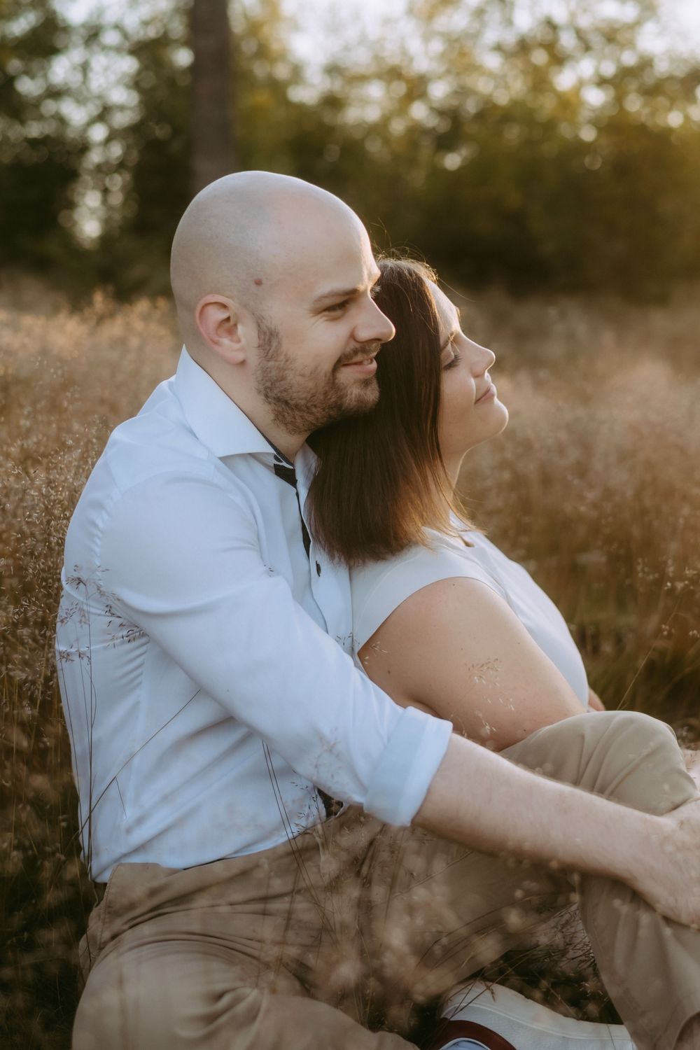 Couple sitting together in sunlit meadow, woman leaning back against man in light blue shirt at dusk.