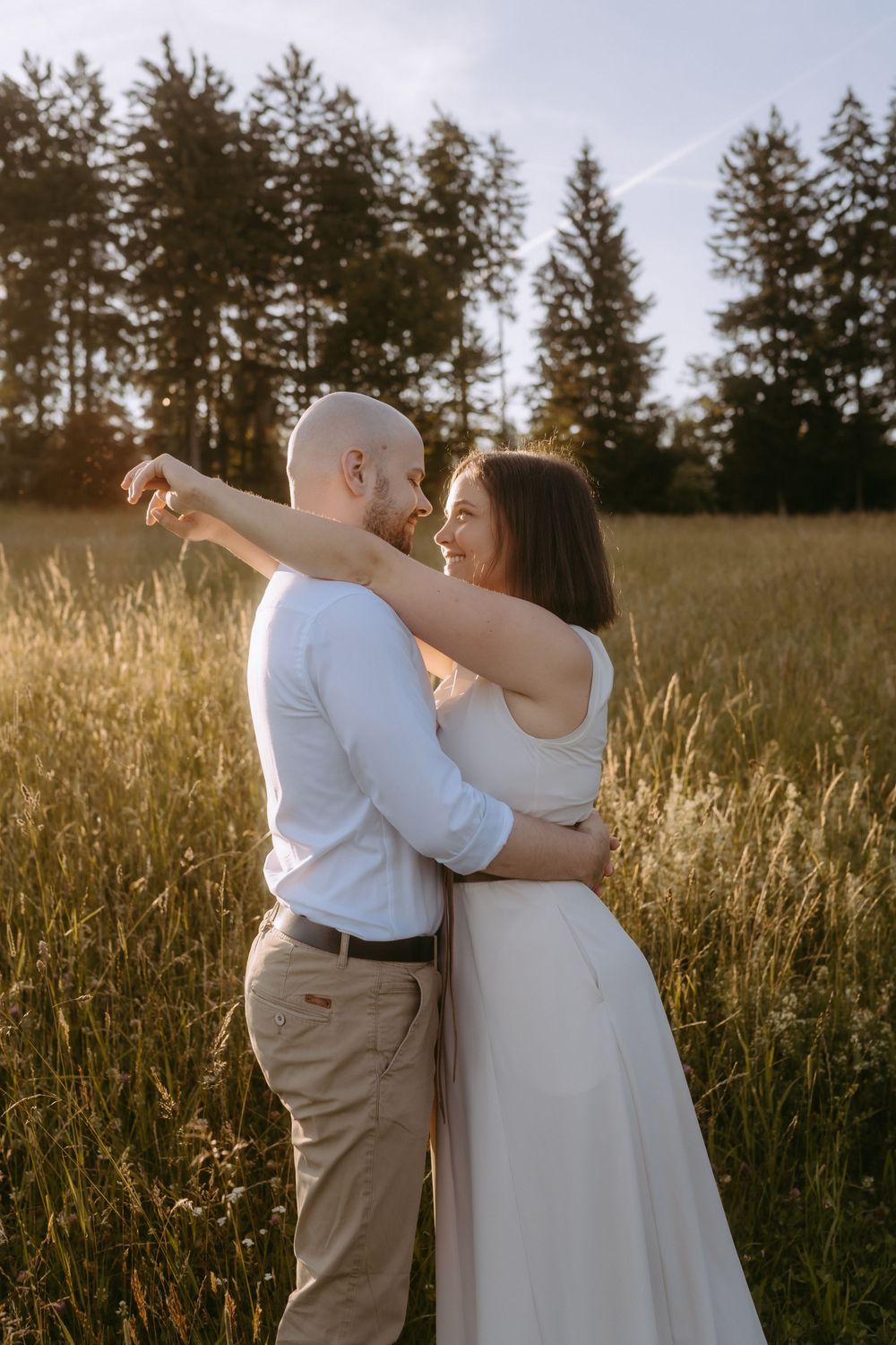 Couple embracing in golden field at sunset, woman in white dress smiling at bald man in white shirt.