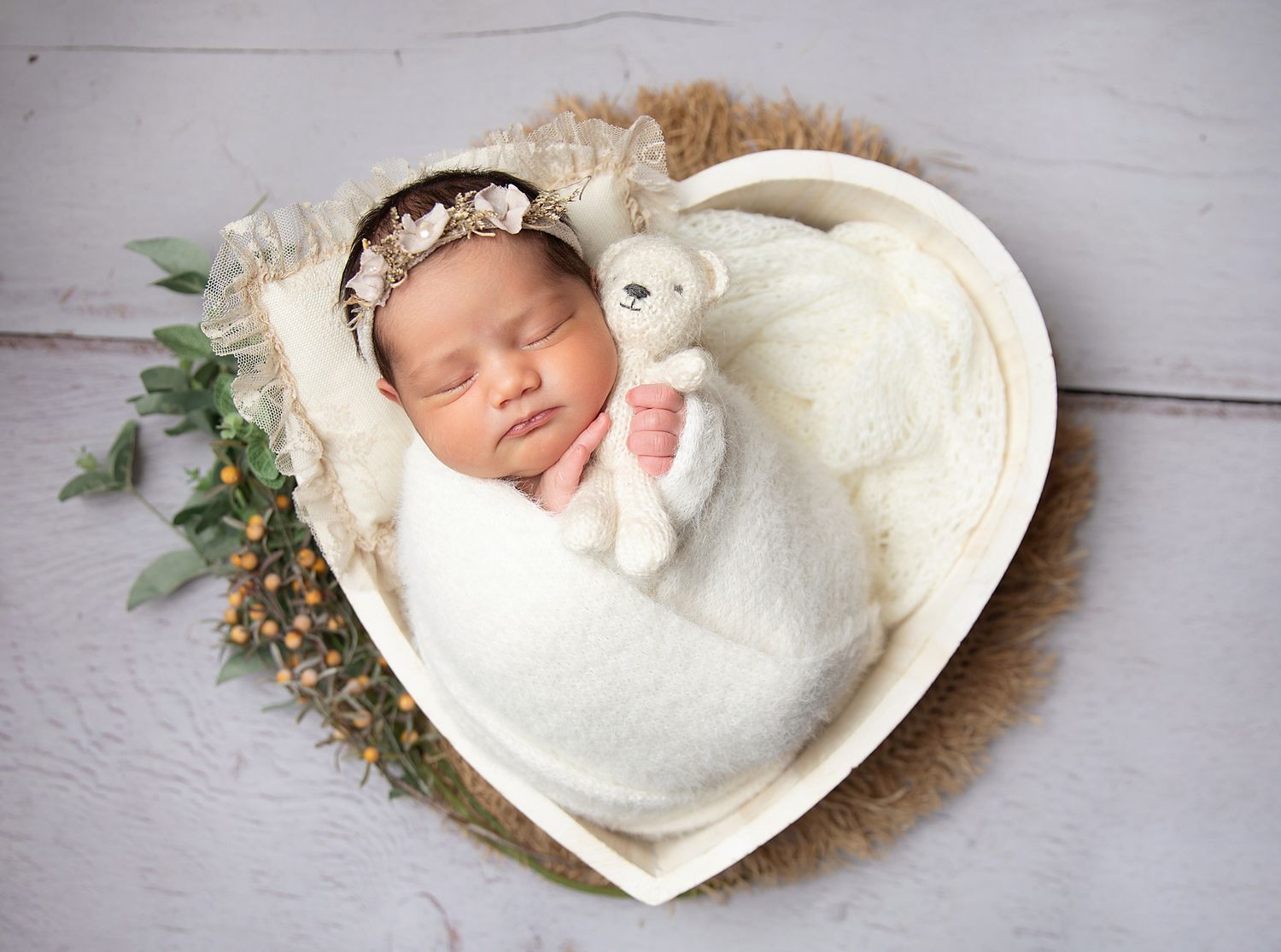 Sleeping newborn baby wrapped in white swaddle holding teddy bear in heart-shaped wooden bowl with floral headband. During a newborn session