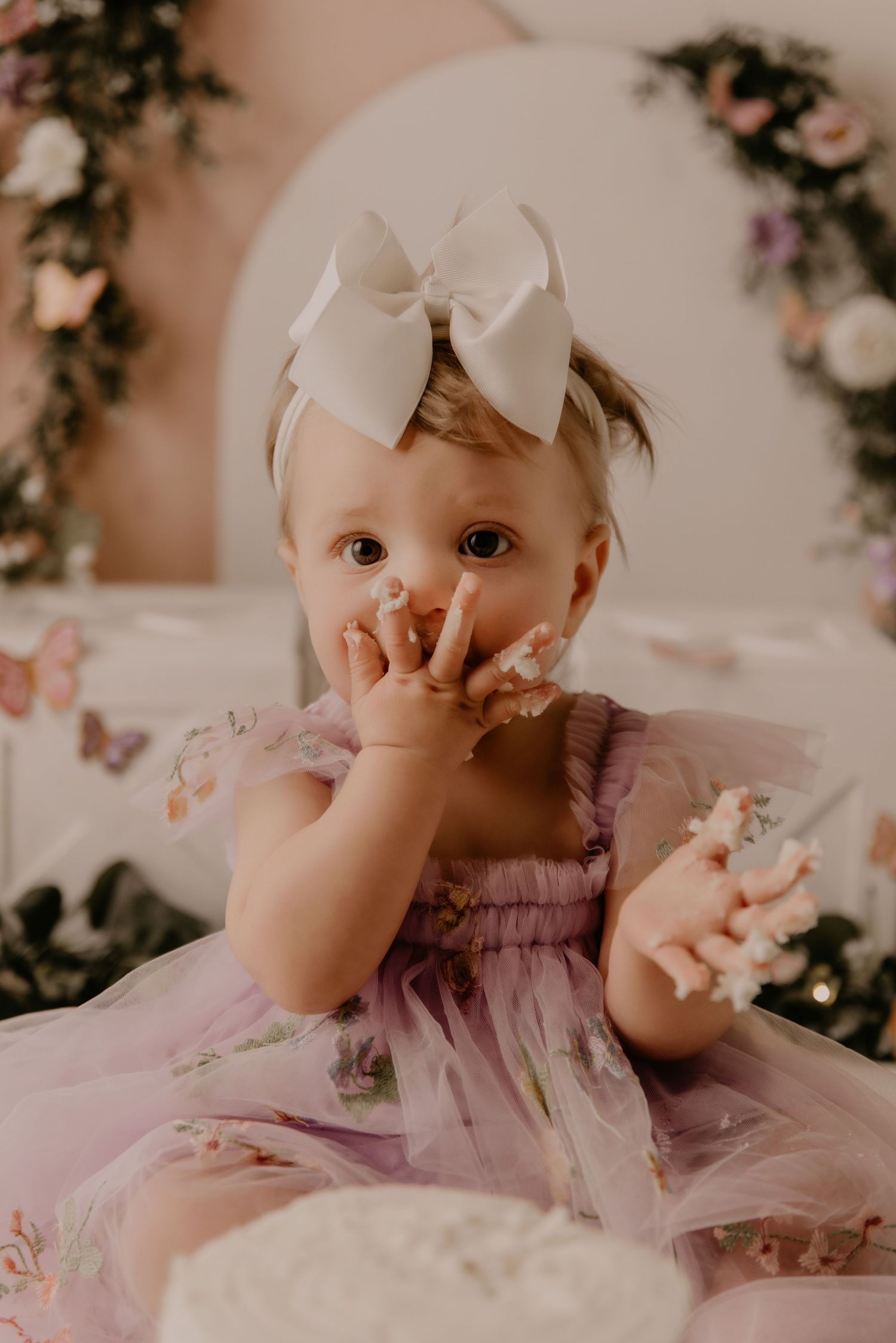 Baby girl in pink floral tutu dress with white bow headband eating birthday cake at first birthday smash cake photoshoot.