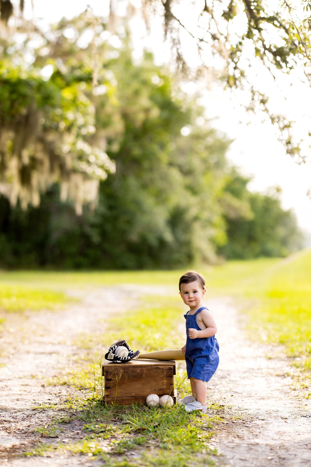 Toddler boy in blue romper standing on dirt path with vintage wooden crate, baseball bat, glove, and balls outdoors.