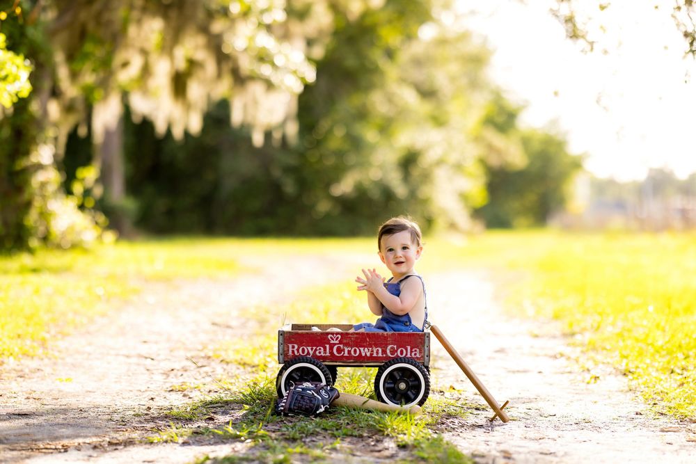 Toddler boy sitting in a red Royal Crown Cola wagon on a sunny tree-lined dirt path.