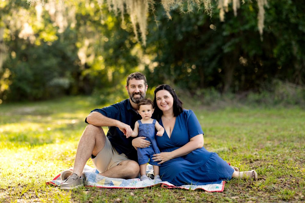 A happy family of three sits on a blanket outdoors, smiling, with lush green trees and Spanish moss in the background.