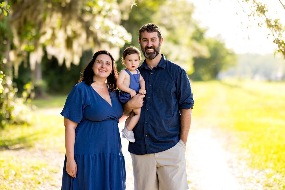 A smiling family of three poses on a sunny tree-lined path, parents in blue outfits holding their toddler.