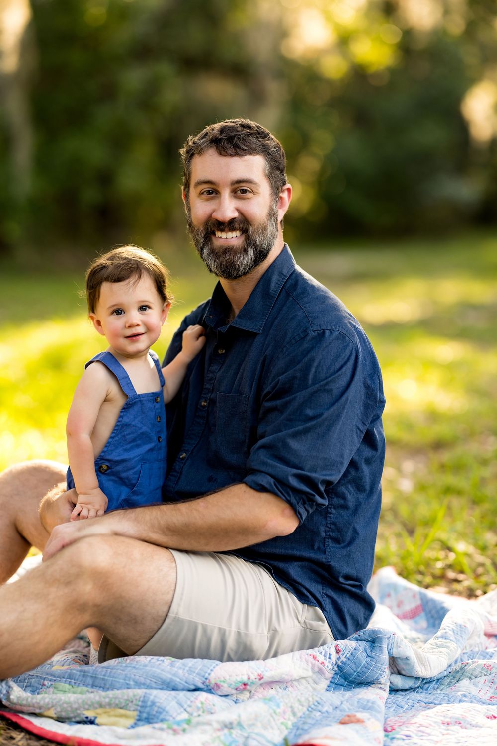 Man with beard sits on quilt in park holding smiling baby in blue overalls, enjoying outdoor family time.