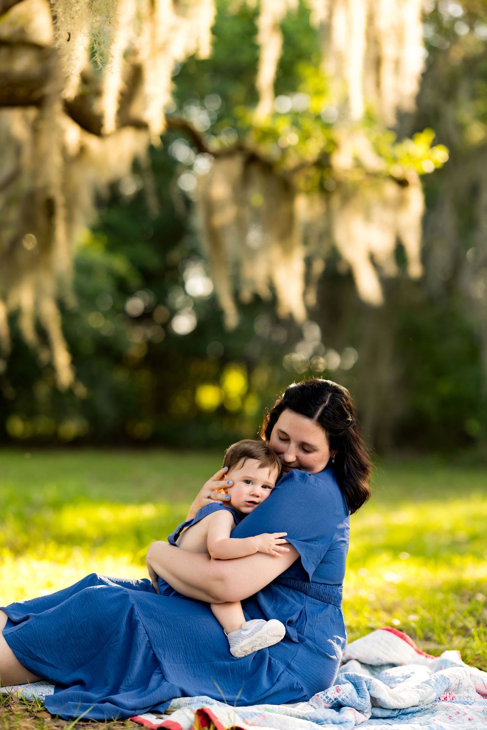 Mother in blue dress tenderly embraces baby on blanket under Spanish moss-draped tree in sunny park.