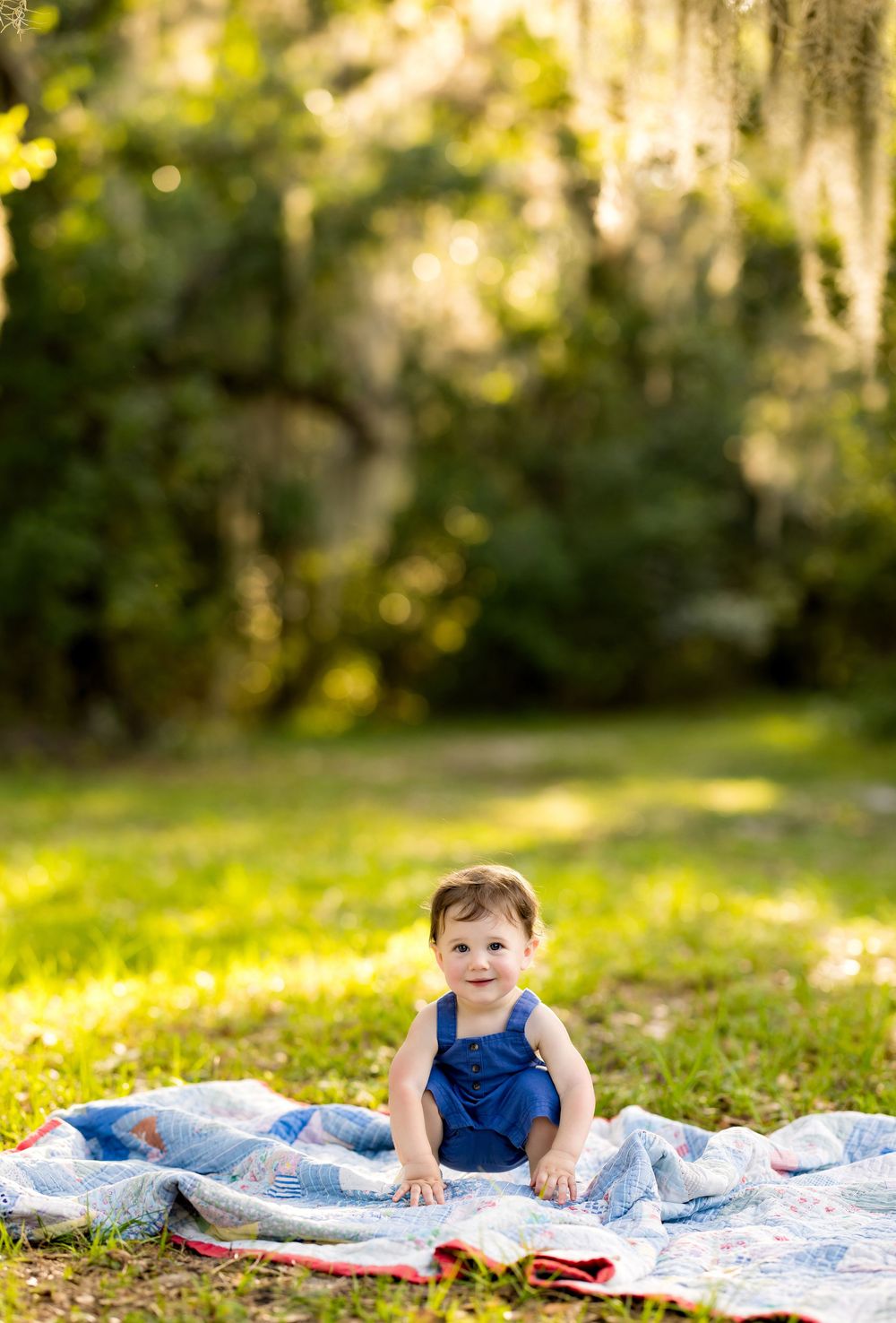 A smiling baby in blue overalls sits on a quilt blanket in a sunny green park with mossy trees.