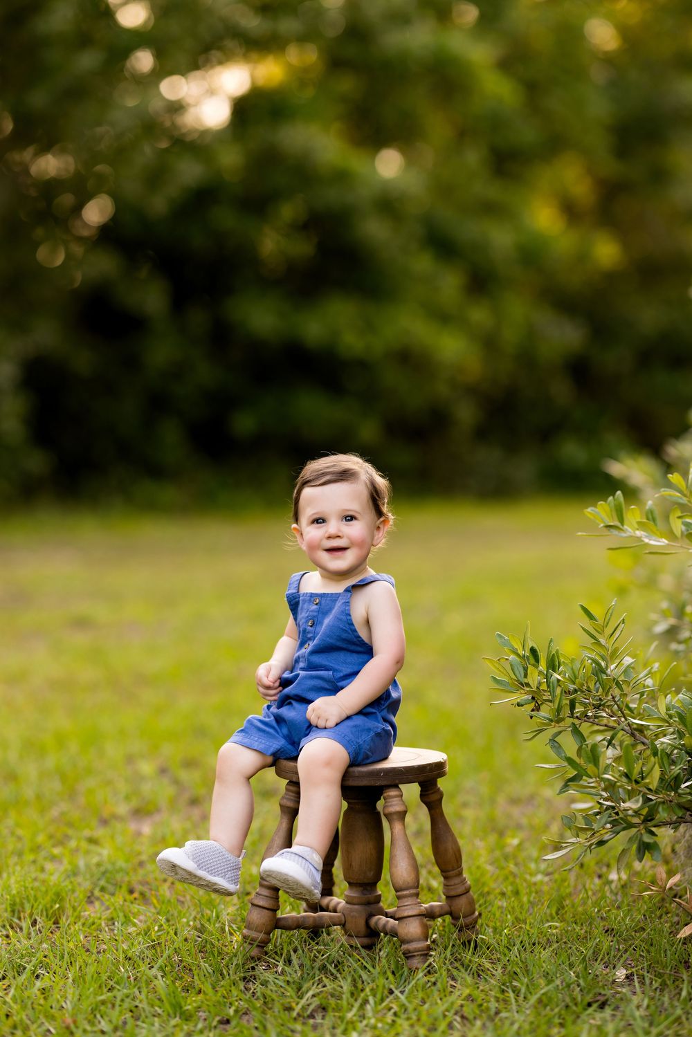 Smiling toddler in blue denim overalls sitting on a wooden stool outdoors on green grass.