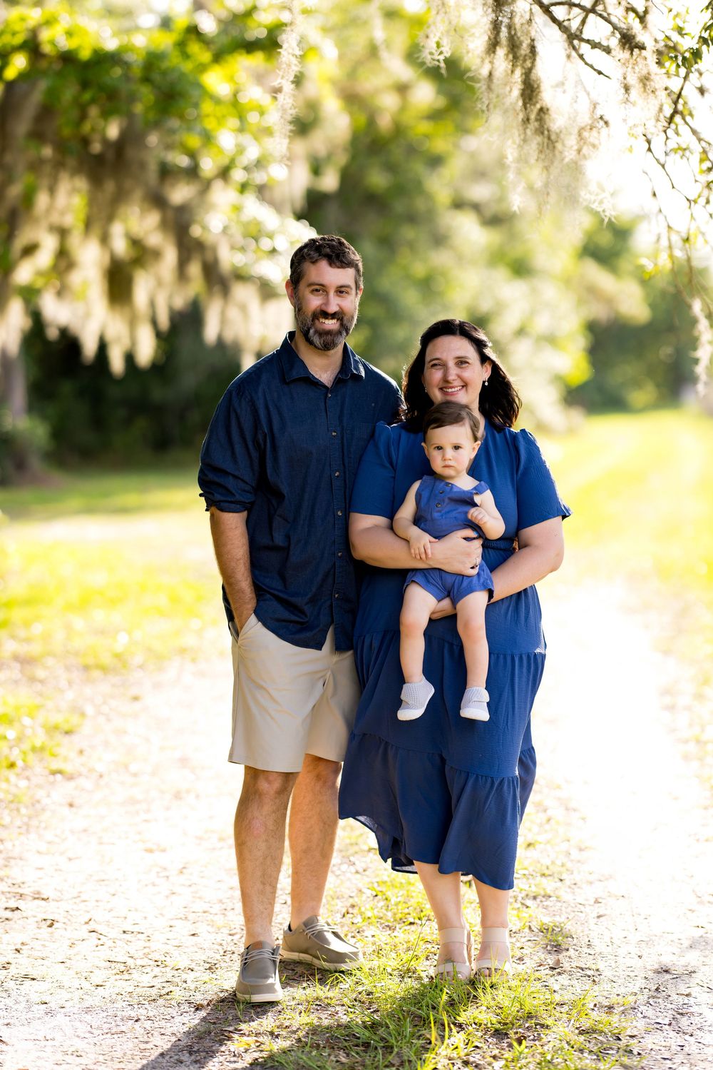 A smiling family of three in matching navy blue outfits poses on a sunlit path surrounded by lush green trees.