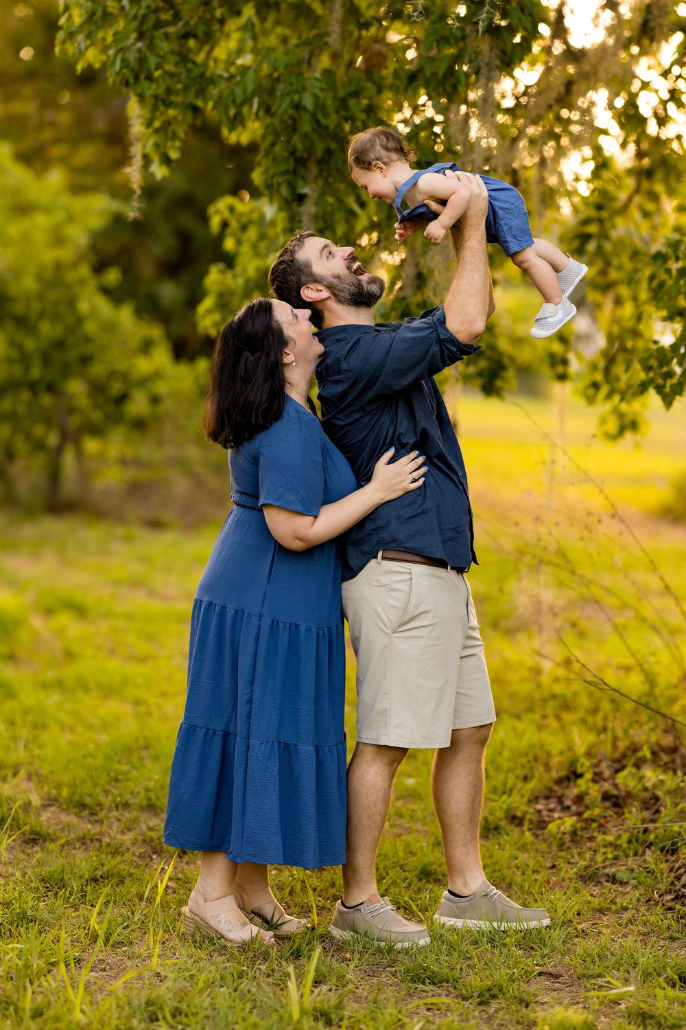 A happy family of three outdoors, parents lifting toddler in blue outfits among lush green trees at golden hour.