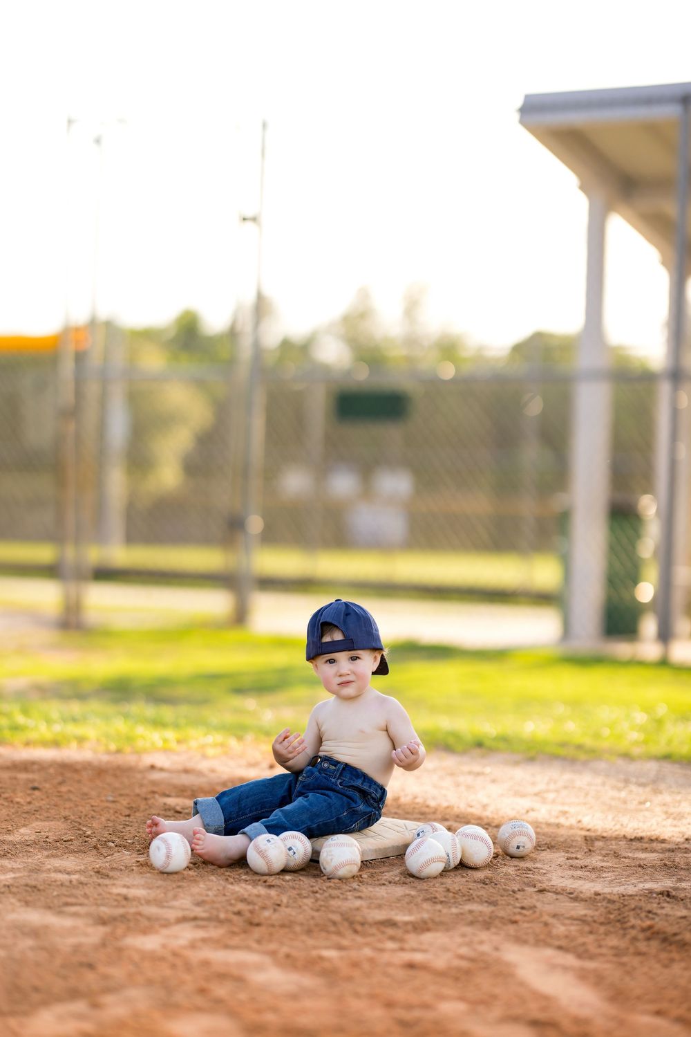Shirtless toddler in blue cap and jeans sits on baseball base surrounded by baseballs on dirt field.