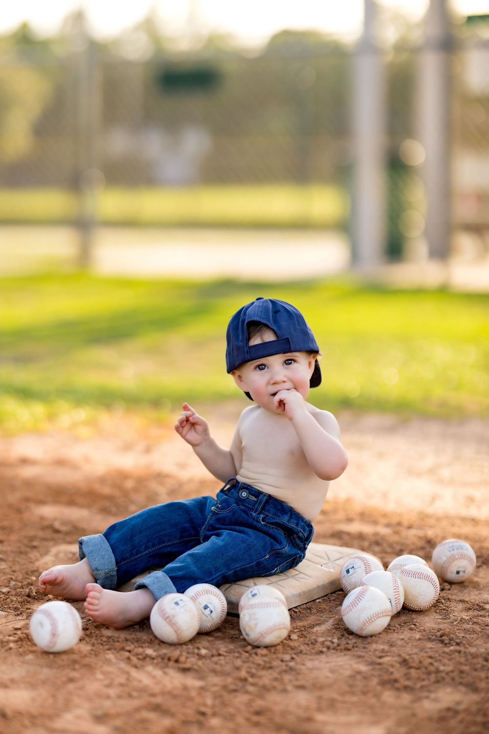Cute baby boy in a baseball cap and jeans sits on a base surrounded by baseballs on a dirt field.
