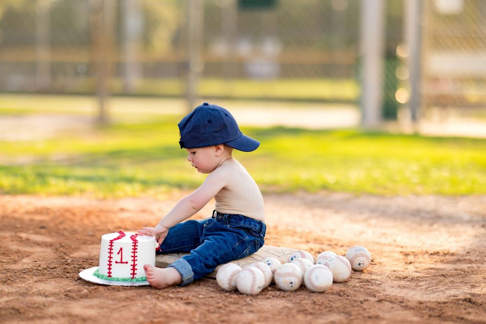 Toddler boy in blue cap sits on baseball diamond dirt beside a number one birthday cake and baseballs.