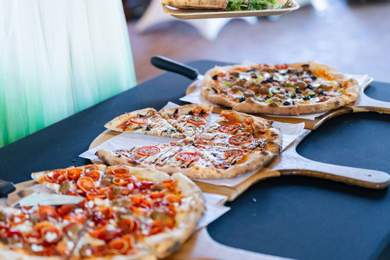 Three assorted pizzas on wooden paddles displayed on a black table at a catered event.