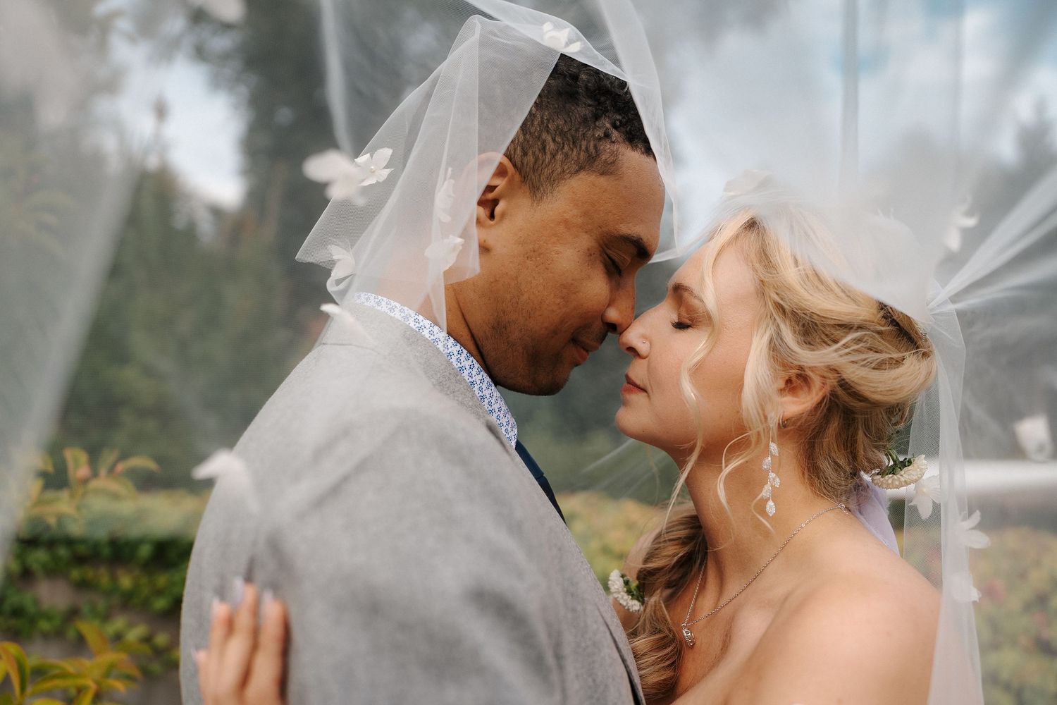 Bride and groom share a romantic moment under a flowing white veil outdoors on their wedding day.
