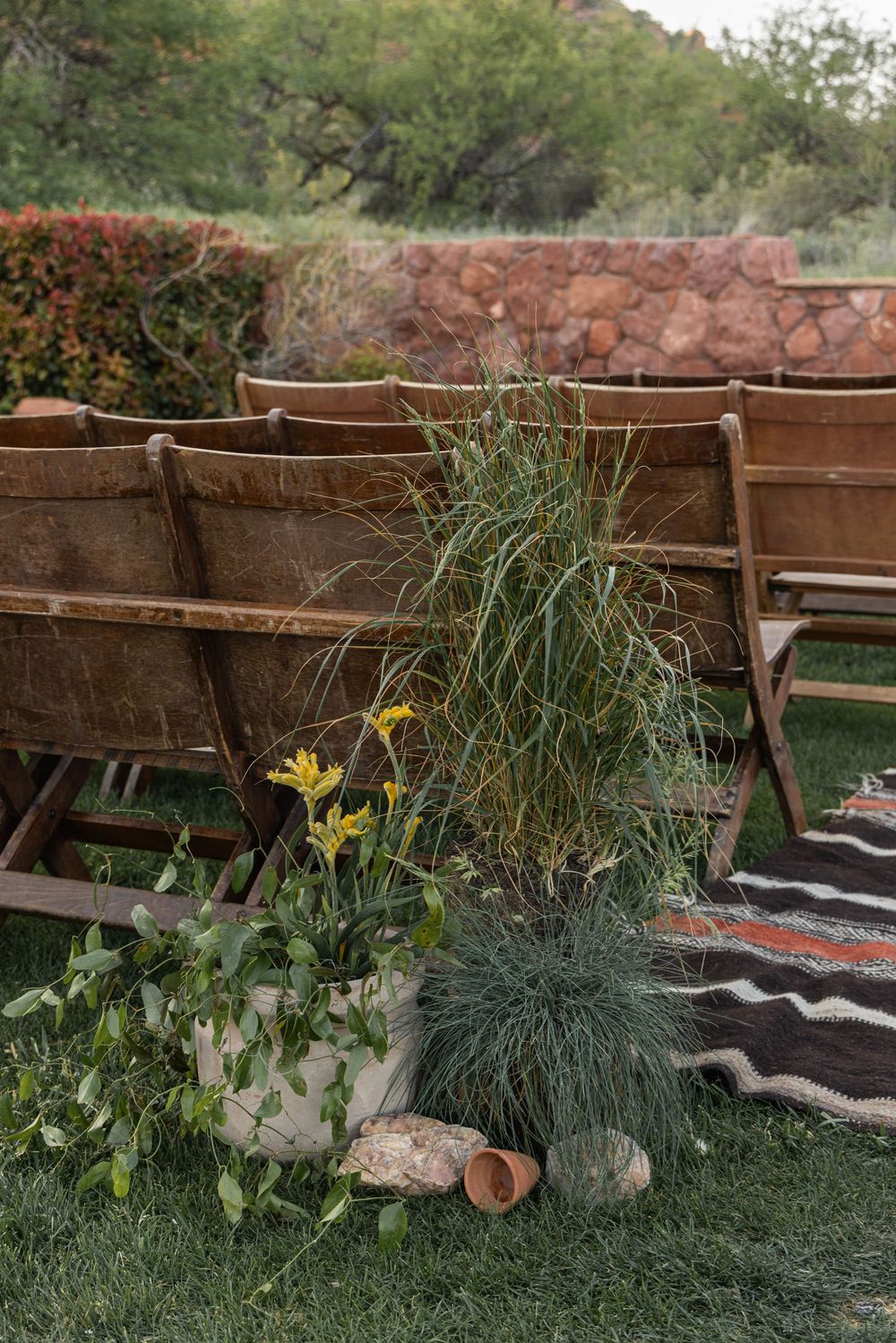 Rustic wooden folding chairs arranged outdoors with potted ornamental grasses and wildflowers on a patterned rug.