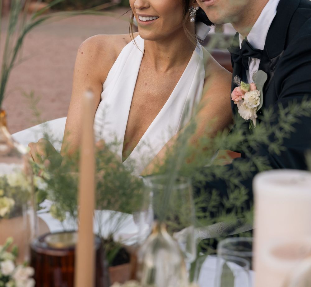 Smiling bride in white halter gown sits beside groom in tuxedo at floral wedding reception table.