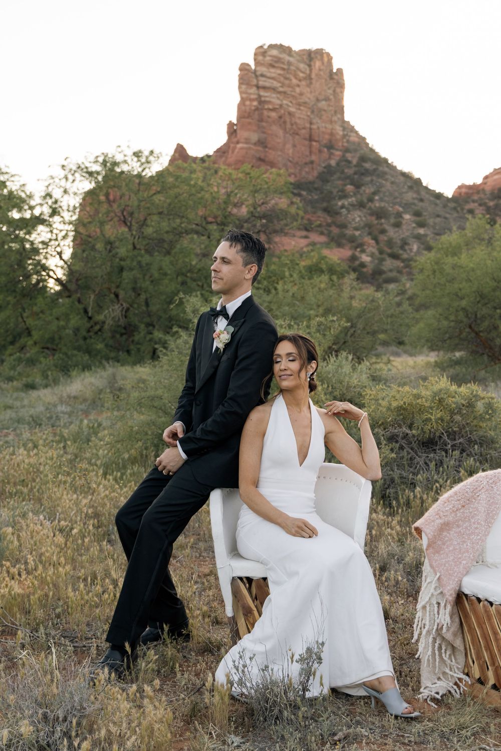 Bride in white halter gown seated in chair with groom in black tuxedo beside her, Sedona red rocks backdrop.