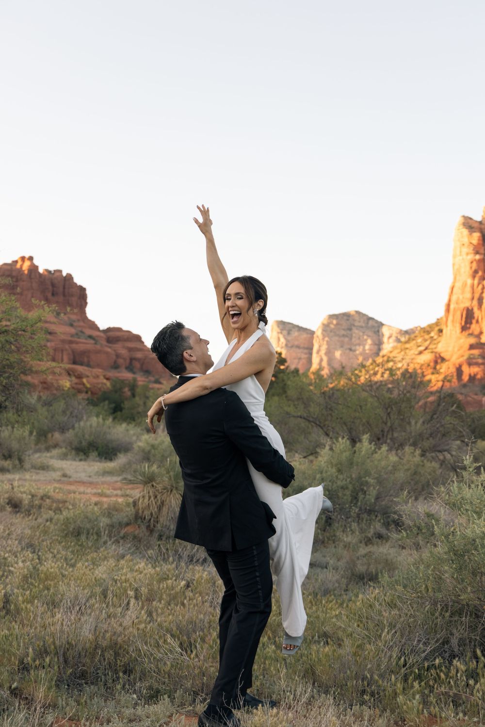 Groom lifts joyful bride in white dress among red rock formations in Sedona, AZ during golden hour wedding photos.