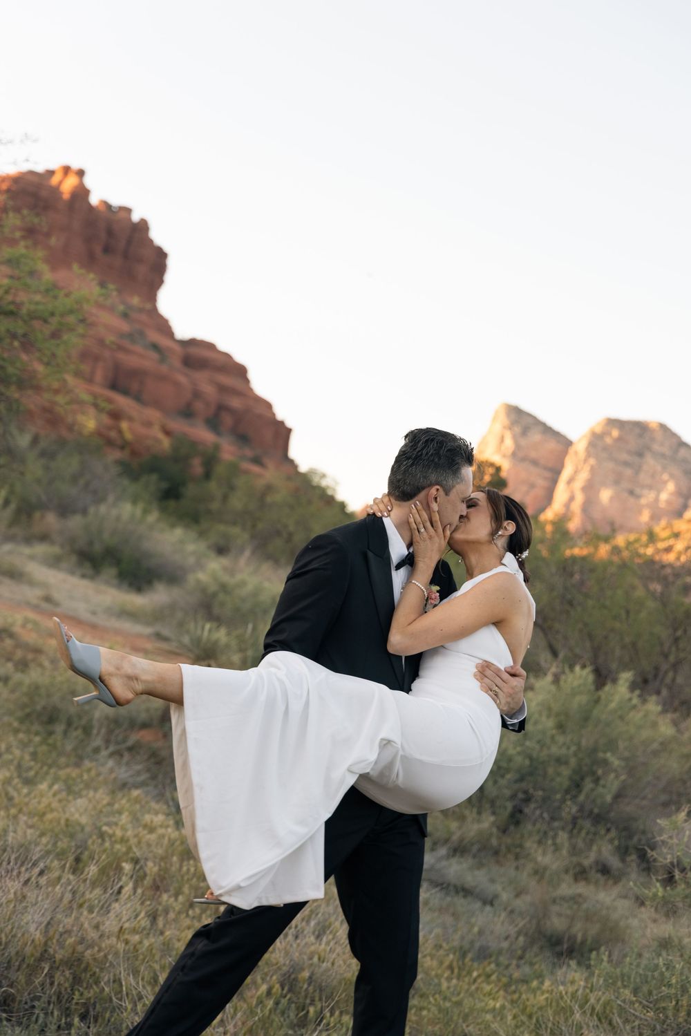 A groom in a black tuxedo lifts his bride in a white dress for a kiss against red rock formations in Sedona.