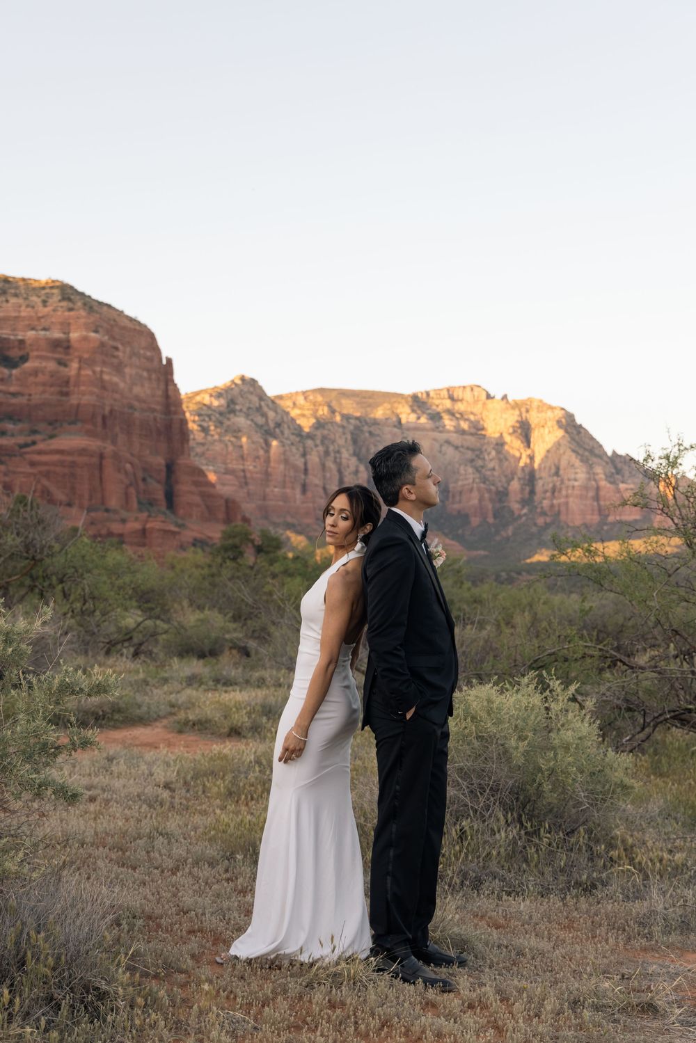 Couple in wedding attire standing back-to-back among desert shrubs with red rock formations in Sedona, Arizona at dusk.