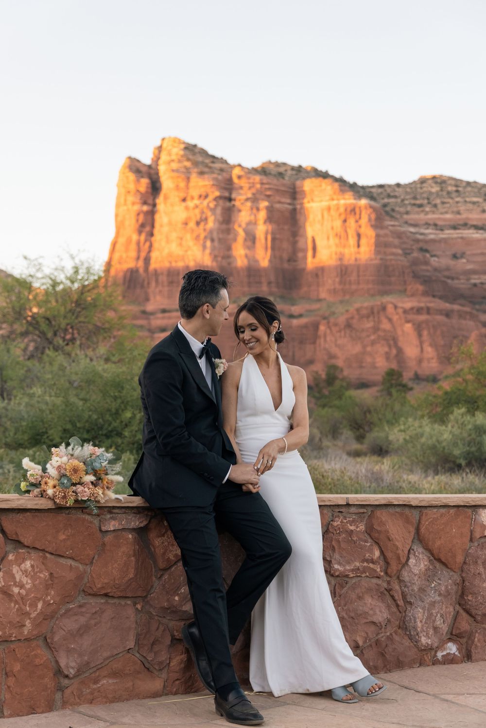Bride and groom share a romantic moment on a stone wall with stunning red rock formations in Sedona, Arizona at sunset.