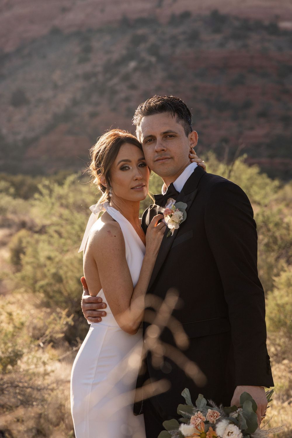 Bride and groom pose in desert landscape with red rock mountains, she in white gown, he in black suit with boutonniere.