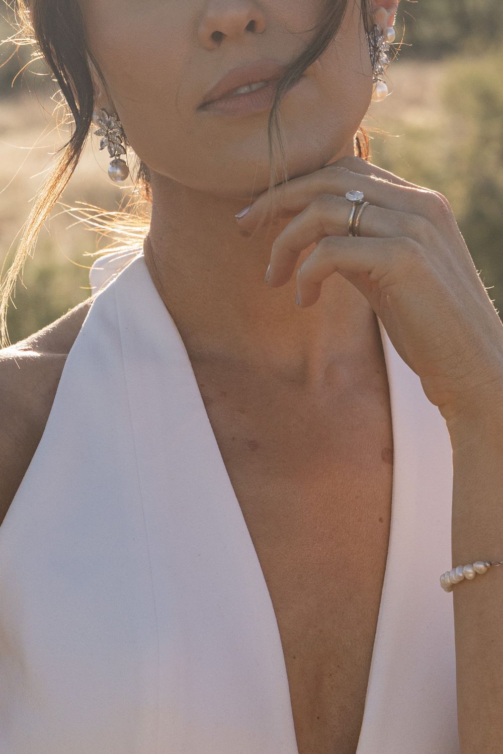 Woman in white halter dress with pearl bracelet, diamond ring, and crystal drop earrings in golden sunlight.