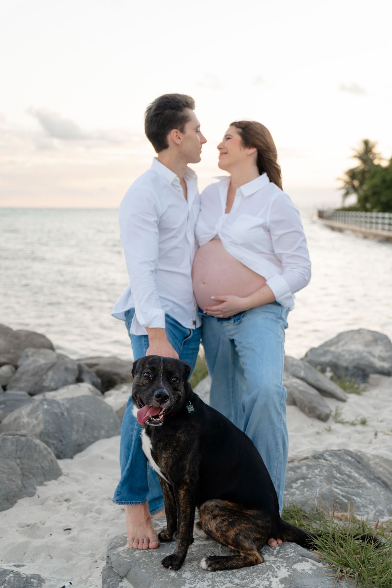 Pregnant couple standing with small white dog wearing blue bandana in park setting.