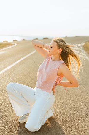Blonde woman in pink top and white wide-leg pants sitting on road with hair blowing in wind.