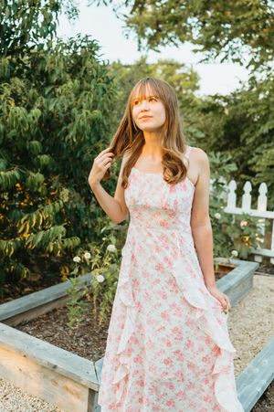 Woman in floral pink maxi dress posing in a garden with white fence and lush green trees in warm sunlight.