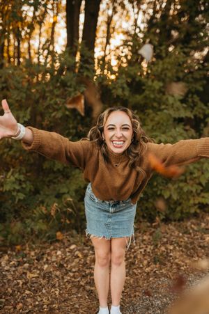 Happy young woman in brown sweater and denim skirt tossing autumn leaves in forest setting.