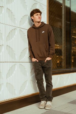 A young man in a brown Nike hoodie and olive pants leans against a decorative white wall.