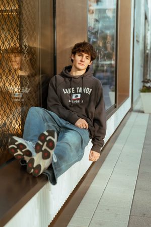 Teen boy in Lake Tahoe hoodie and jeans sitting on storefront ledge in warm sunlight.