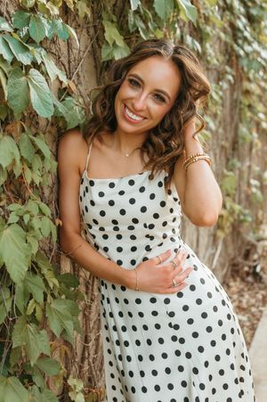 Smiling woman in white polka dot sundress leaning against tree surrounded by lush green ivy.