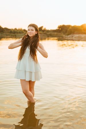 Young woman in white tiered dress standing in shallow water during golden sunset.