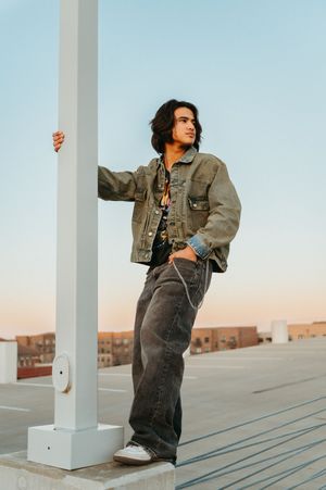Young man in denim jacket and wide-leg jeans leaning on pole on rooftop at sunset.
