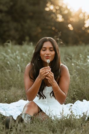 Young woman in white dress sitting in a field, eyes closed, blowing a dandelion at golden hour.