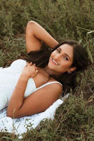 Smiling brunette woman in white dress lying in green grass field looking up at camera.