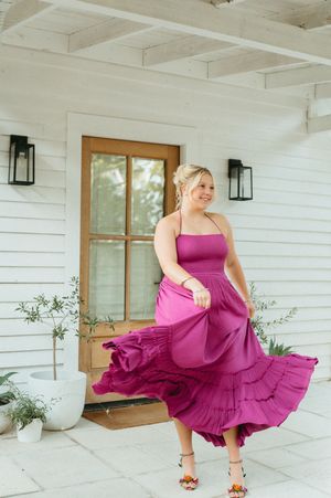 Woman twirling in a vibrant magenta midi dress with tiered skirt, smiling outdoors near a white farmhouse door.