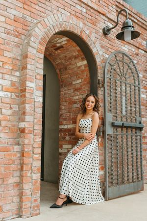 A smiling woman in a white polka dot maxi dress poses in a brick archway doorway.