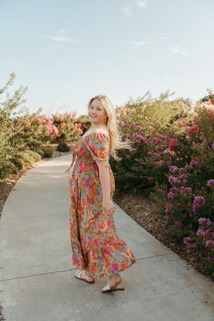 Blonde woman in colorful floral maxi dress walking on garden path surrounded by pink blooming flowers.