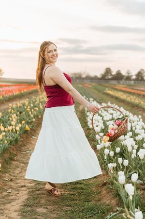 Redhead woman in red top and white tiered skirt holding flower basket in tulip field at sunset.