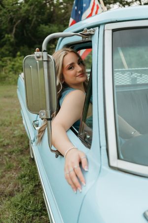 Smiling blonde woman leaning out of a vintage light blue truck window with an American flag inside.