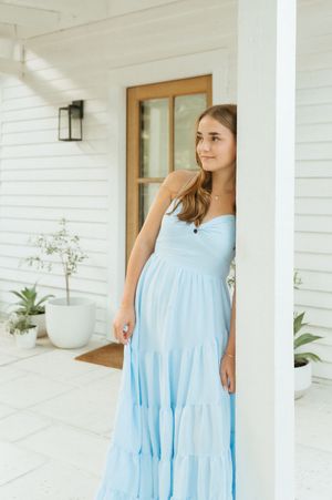 Young woman in a light blue strapless maxi dress posing by a white porch column of a farmhouse.
