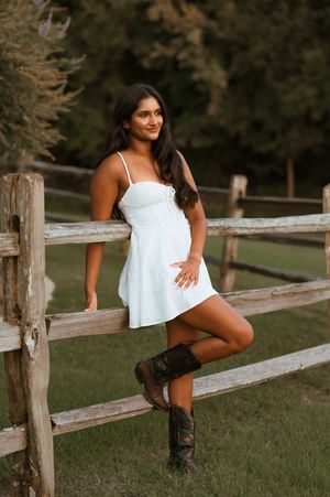 Smiling woman in white sundress and cowboy boots posing against a rustic wooden fence outdoors.