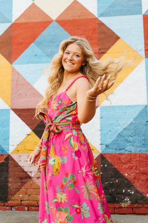 Smiling blonde woman in pink tropical floral dress posing in front of colorful geometric mural wall.