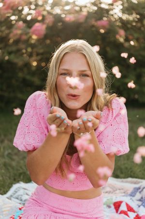 Blonde girl in pink outfit blowing flower petals outdoors in garden setting.